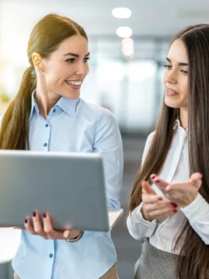 **Descripción SEO:**

En esta imagen se puede ver a dos mujeres profesionales en un entorno de oficina moderno, interactuando mientras miran una computadora portátil. Una de ellas, con una camisa azul, sonríe mientras sostiene el dispositivo, mostrando un ambiente de colaboración y trabajo en equipo. La otra mujer, vestida con una blusa blanca, gesticula mientras explica algo, lo que sugiere una conversación activa y productiva. Este tipo de interacciones son comunes en el ámbito laboral
