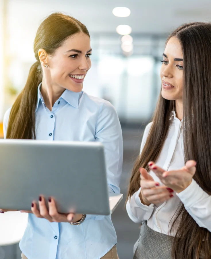 **Descripción SEO:**

En esta imagen se puede ver a dos mujeres profesionales en un entorno de oficina moderno, interactuando mientras miran una computadora portátil. Una de ellas, con una camisa azul, sonríe mientras sostiene el dispositivo, mostrando un ambiente de colaboración y trabajo en equipo. La otra mujer, vestida con una blusa blanca, gesticula mientras explica algo, lo que sugiere una conversación activa y productiva. Este tipo de interacciones son comunes en el ámbito laboral