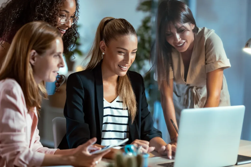 **Descripción de la imagen:**

Grupo de mujeres profesionales colaborando en un entorno de oficina moderno. Una de ellas, sentada frente a una computadora portátil, sonríe mientras revisa información en la pantalla, mientras que las otras tres observan con interés. La escena refleja un ambiente de trabajo colaborativo y creativo, ideal para ilustrar temas de trabajo en equipo, empoderamiento femenino y tecnología en el ámbito laboral. Este tipo de imágenes puede ser útil para atraer a audiencias interesadas