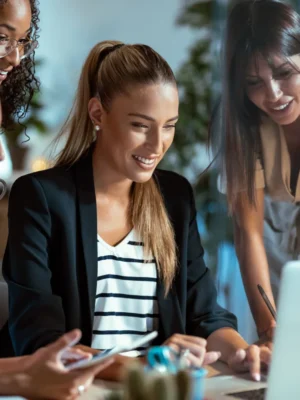 **Descripción de la imagen:**

Grupo de mujeres profesionales colaborando en un entorno de oficina moderno. Una de ellas, sentada frente a una computadora portátil, sonríe mientras revisa información en la pantalla, mientras que las otras tres observan con interés. La escena refleja un ambiente de trabajo colaborativo y creativo, ideal para ilustrar temas de trabajo en equipo, empoderamiento femenino y tecnología en el ámbito laboral. Este tipo de imágenes puede ser útil para atraer a audiencias interesadas