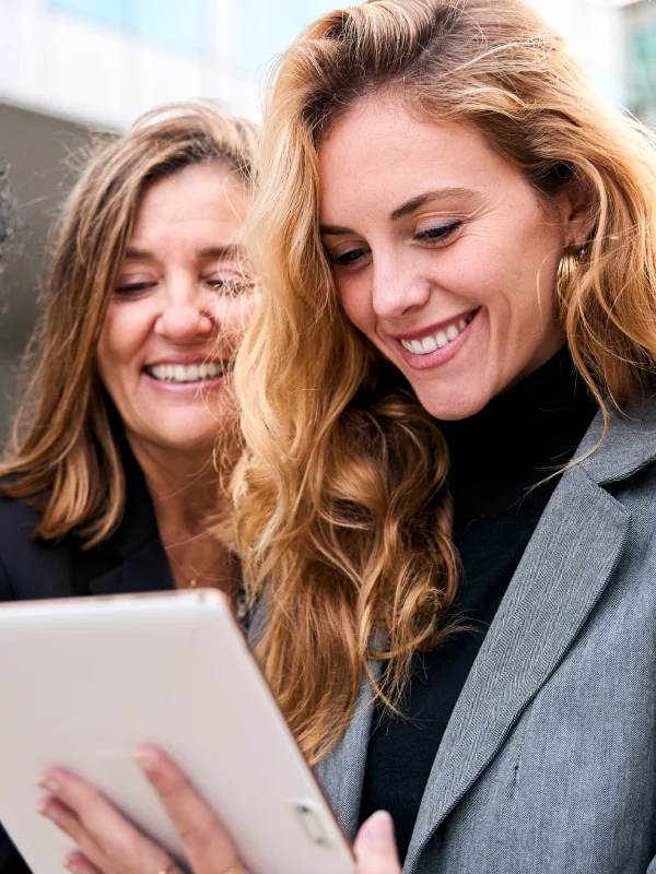**Descripción SEO:**

"Dos mujeres sonrientes revisando contenido en una tablet en un entorno urbano moderno. La imagen captura un momento de conexión y colaboración, destacando la importancia de la tecnología en la comunicación actual. Ideal para ilustrar temas relacionados con trabajo en equipo, innovación y el uso de dispositivos digitales en la vida diaria."