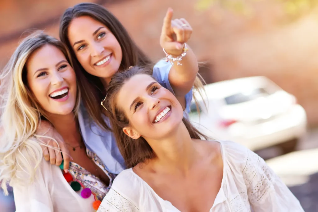 Tres amigas sonrientes disfrutan de un momento divertido al aire libre. La imagen captura la alegría y la complicidad entre ellas, resaltando la importancia de la amistad. Con un fondo desenfocado que sugiere un ambiente urbano, esta fotografía transmite un mensaje positivo y vibrante, ideal para contenido relacionado con la amistad, eventos sociales o actividades al aire libre. La luz natural realza sus sonrisas, haciendo de esta imagen una excelente opción para atraer a un público que busca inspiración en